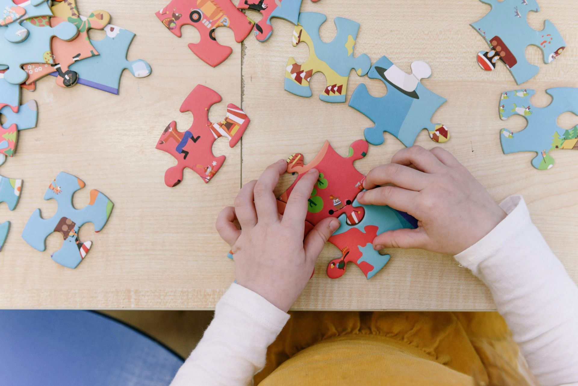 A child’s hands carefully assemble colorful puzzle pieces on a light wooden table.
