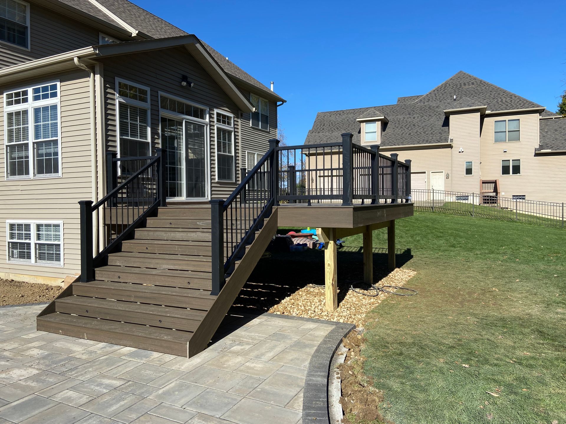 Brown deck and stairs with black railing attached to a beige house, next to a paved pathway and grassy hill.