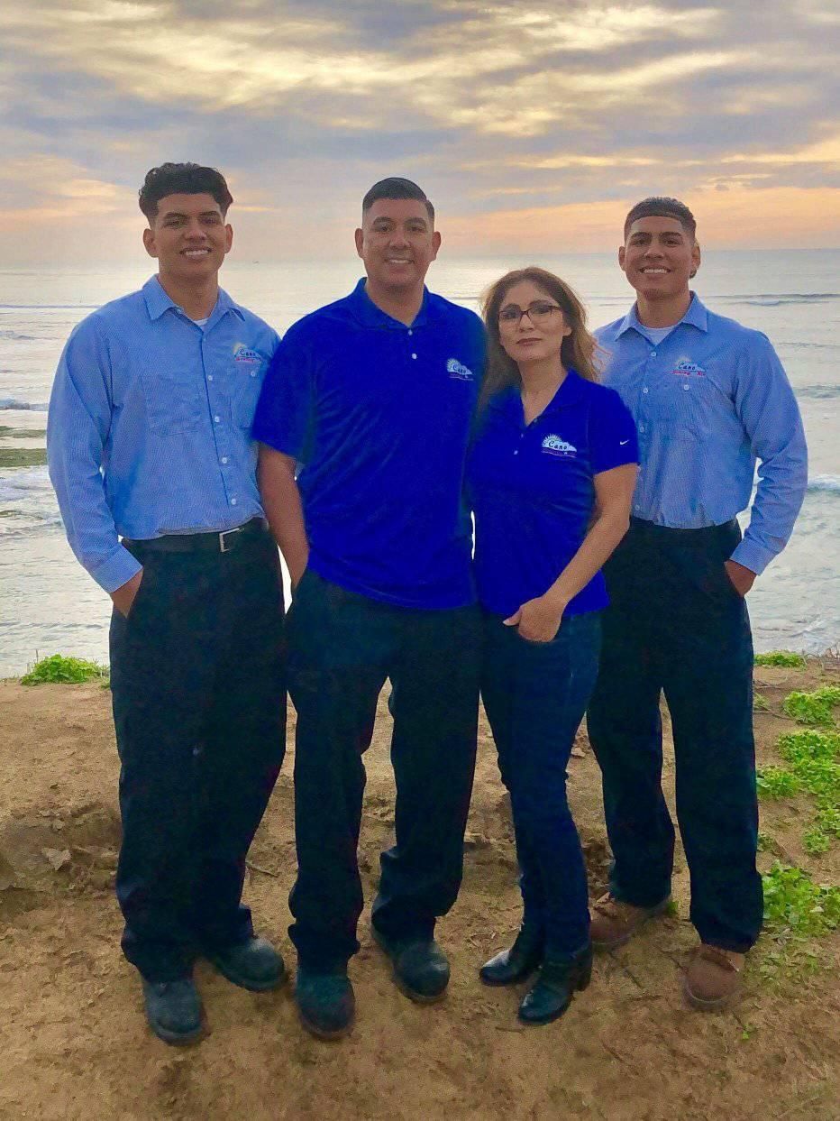 Four people in blue shirts and dark pants pose outdoors near the ocean.