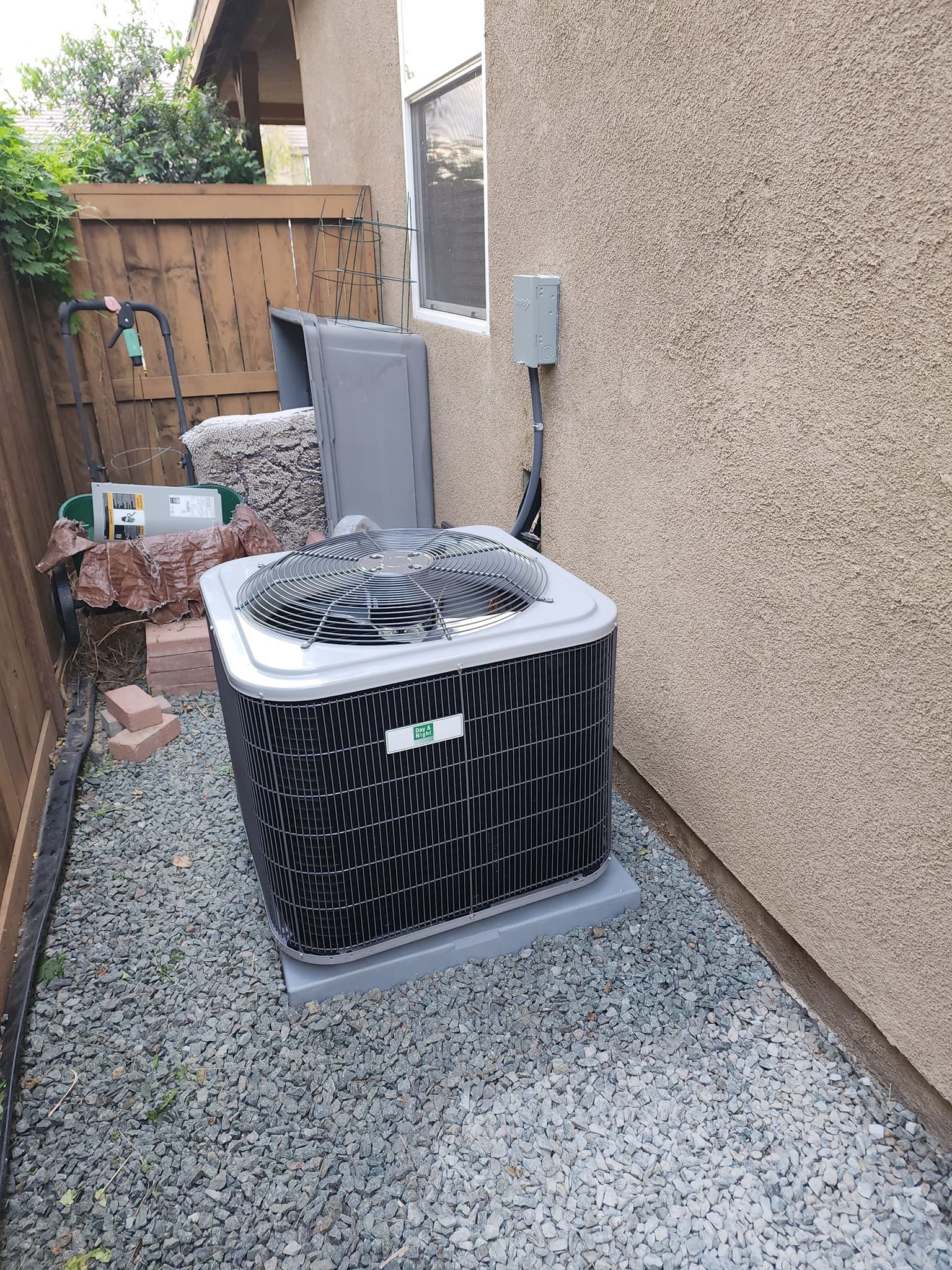AC unit outside house on gravel, next to a wall and fence.
