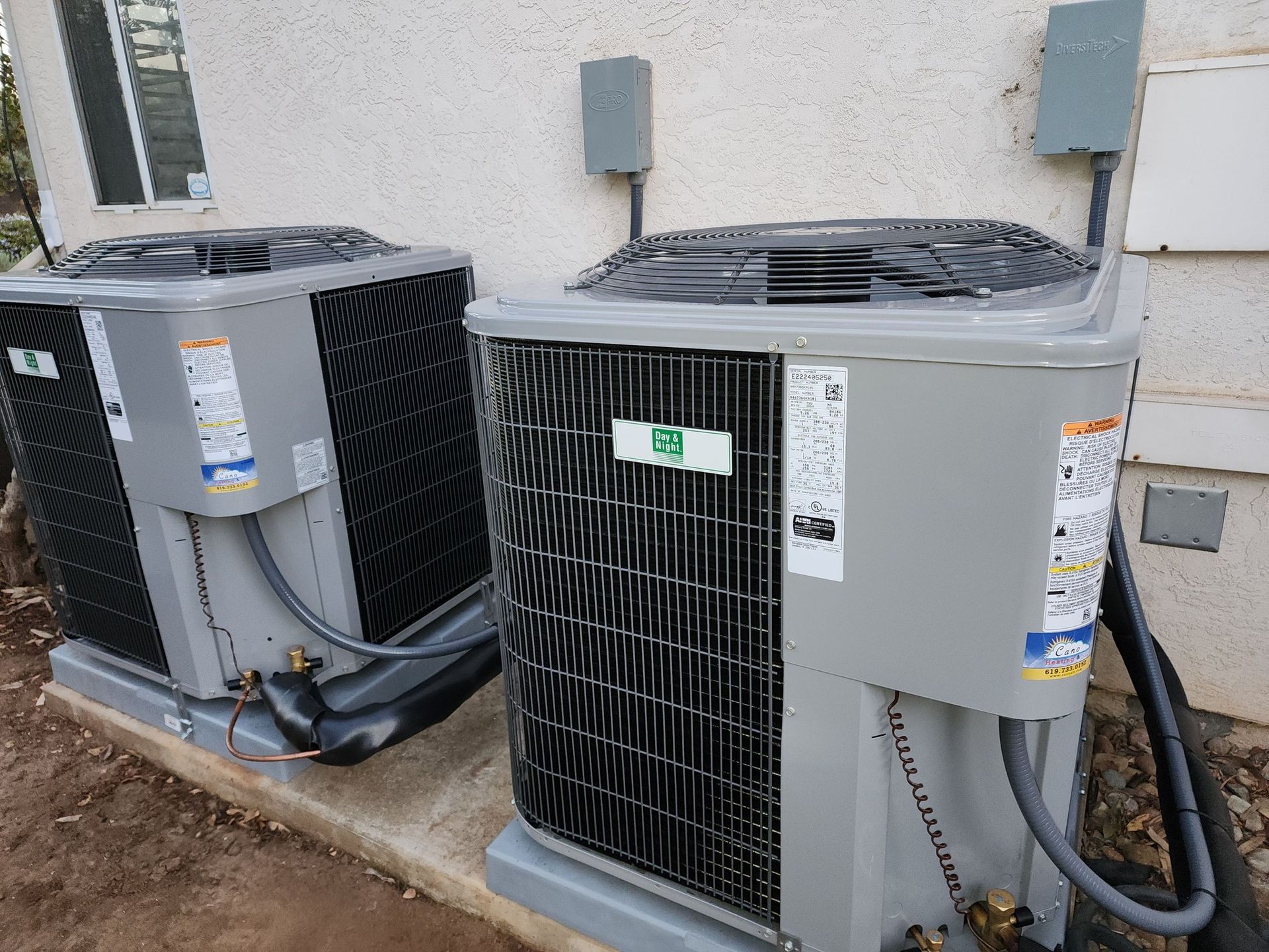Two air conditioning units side-by-side against a beige wall, with electrical boxes above them.