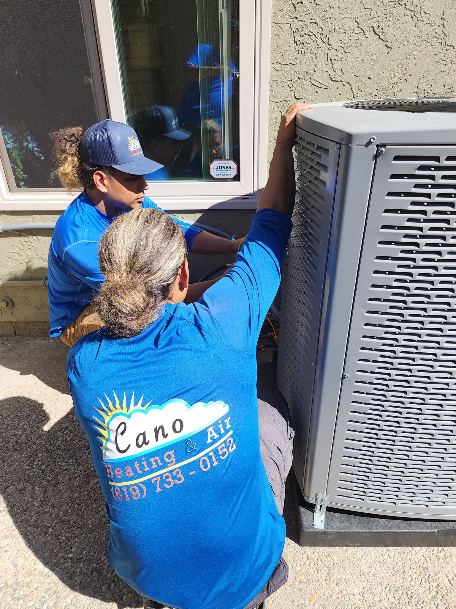 Two people in blue shirts installing an AC unit outside a building.