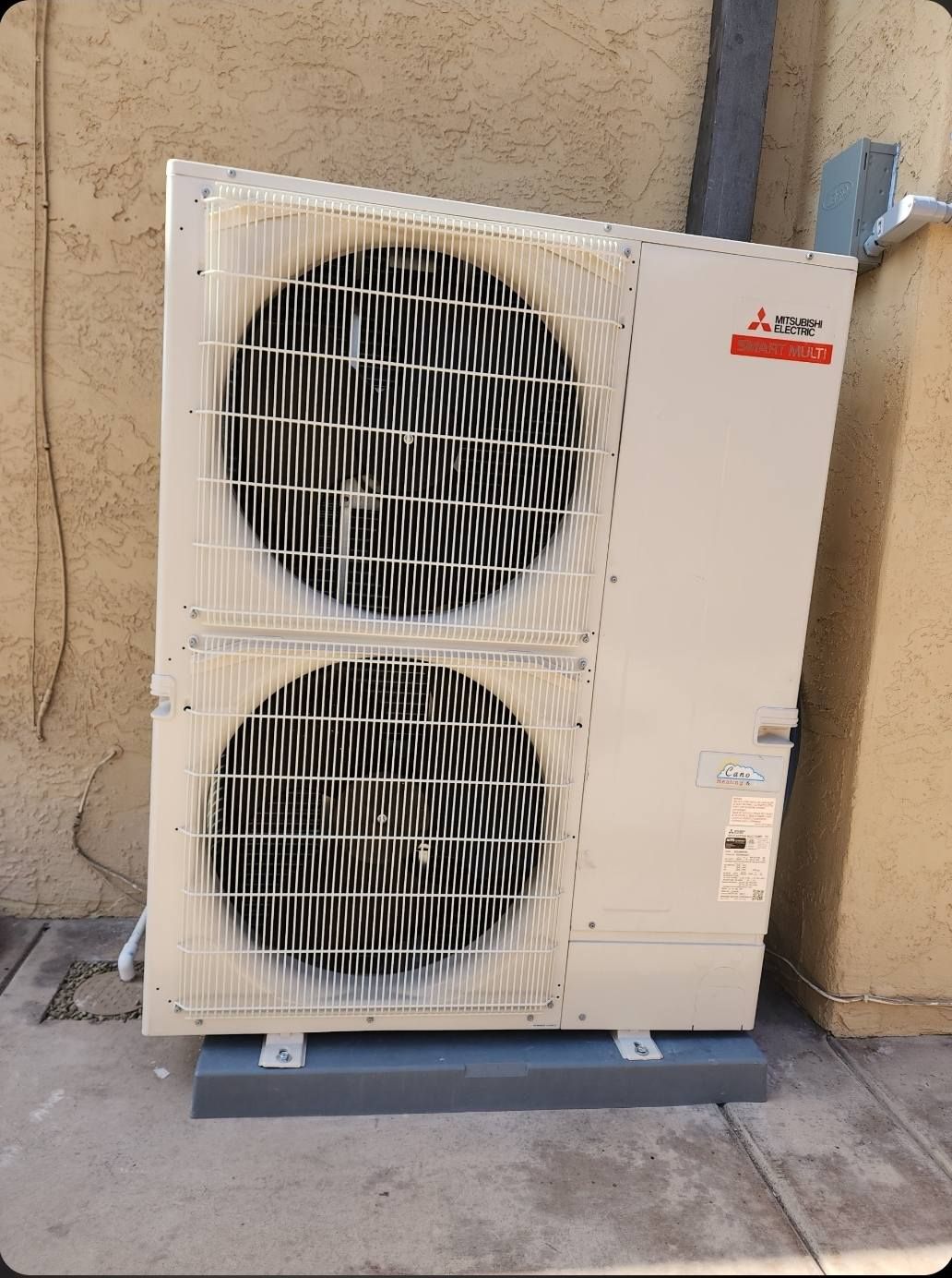 Mitsubishi heat pump unit, beige, with two large fan vents, mounted on a gray base.
