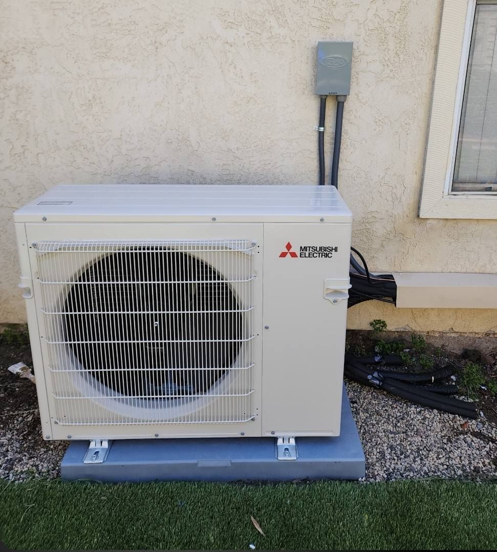 Mitsubishi heat pump on a gray pad next to a beige wall with electrical conduit.