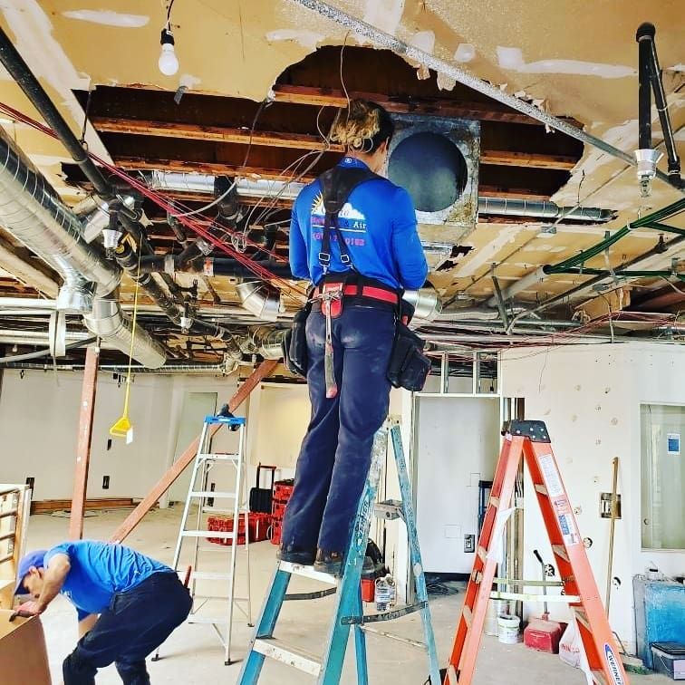Two people working on a ceiling with exposed wires and ductwork. One stands on a ladder, the other works below.