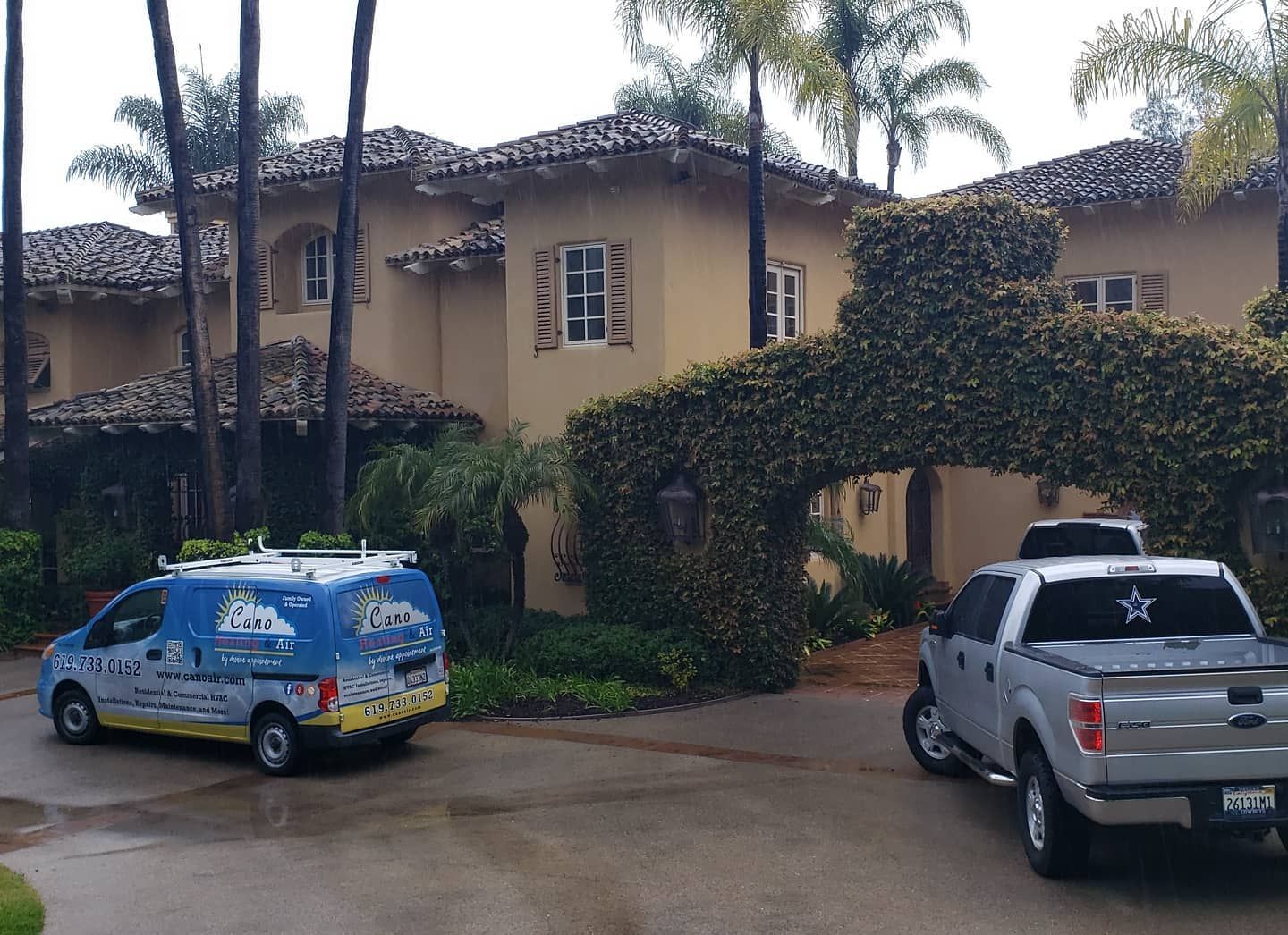 A tan stucco house with a tile roof, ivy archway, and parked service van and truck.