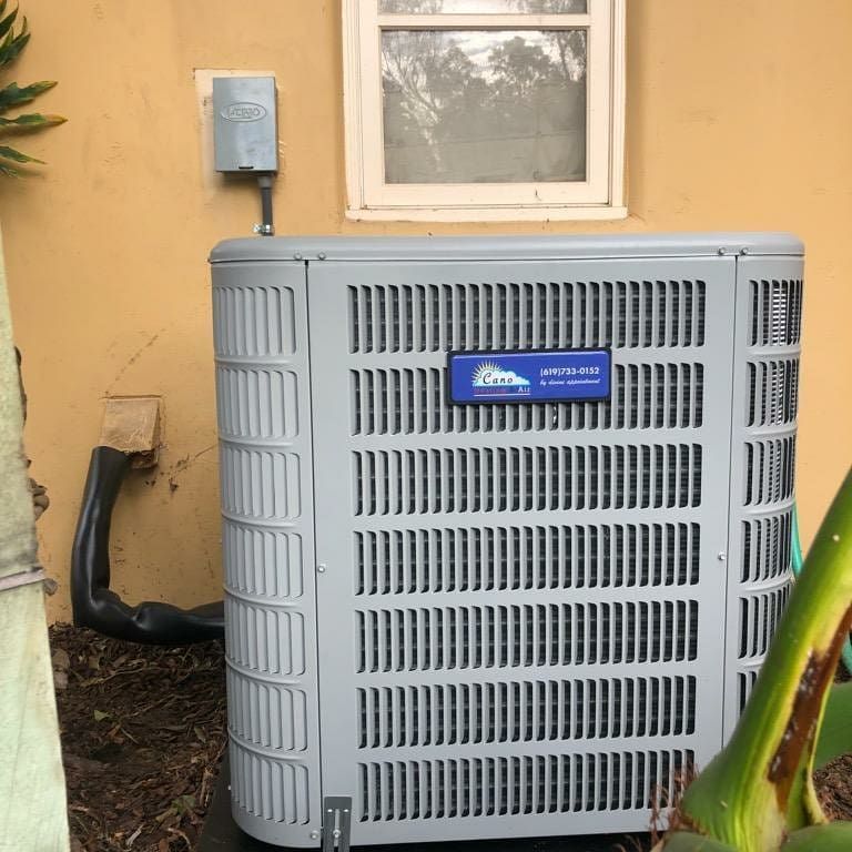 Gray air conditioning unit outside a tan building, with a window and electrical box visible.