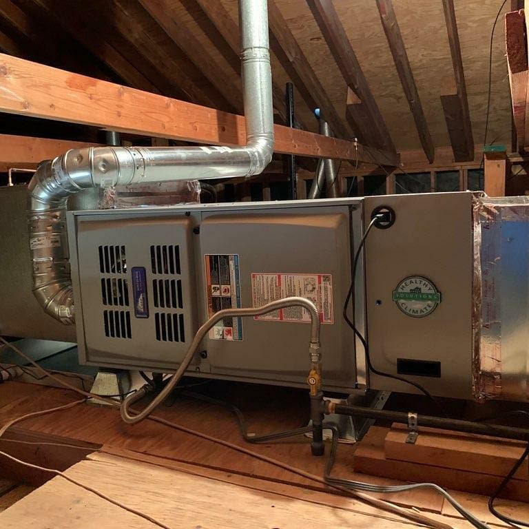 Furnace in an attic. Ductwork is connected to the top and side. Gray metal box with a label. Wooden beams overhead.