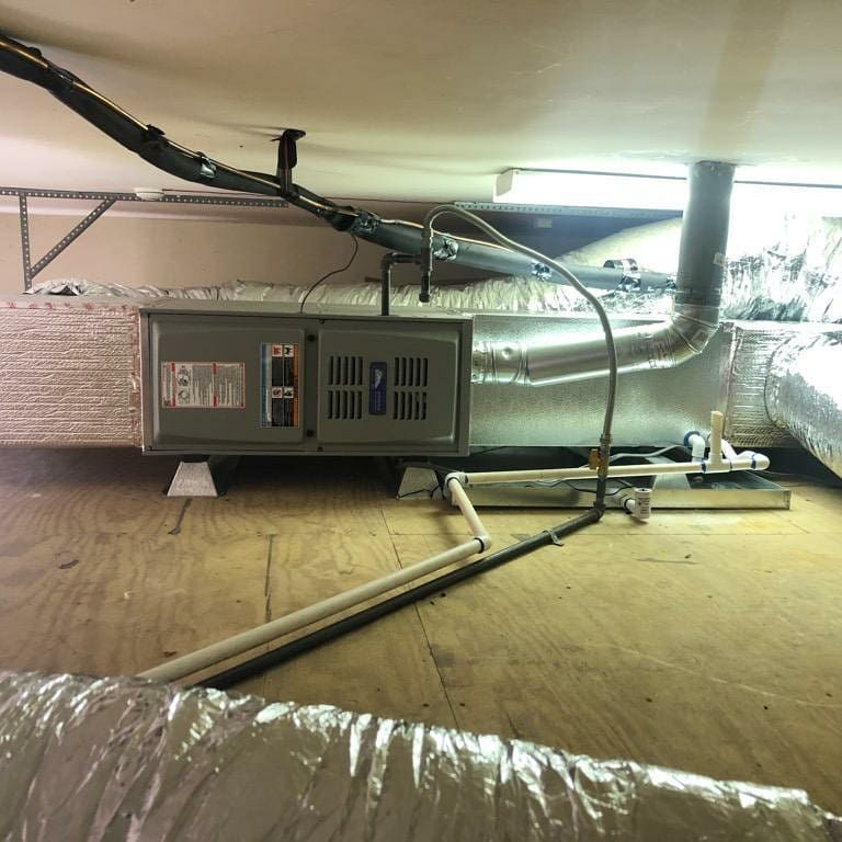 HVAC unit in an attic with ductwork and piping. The unit is silver and gray, mounted on wood.