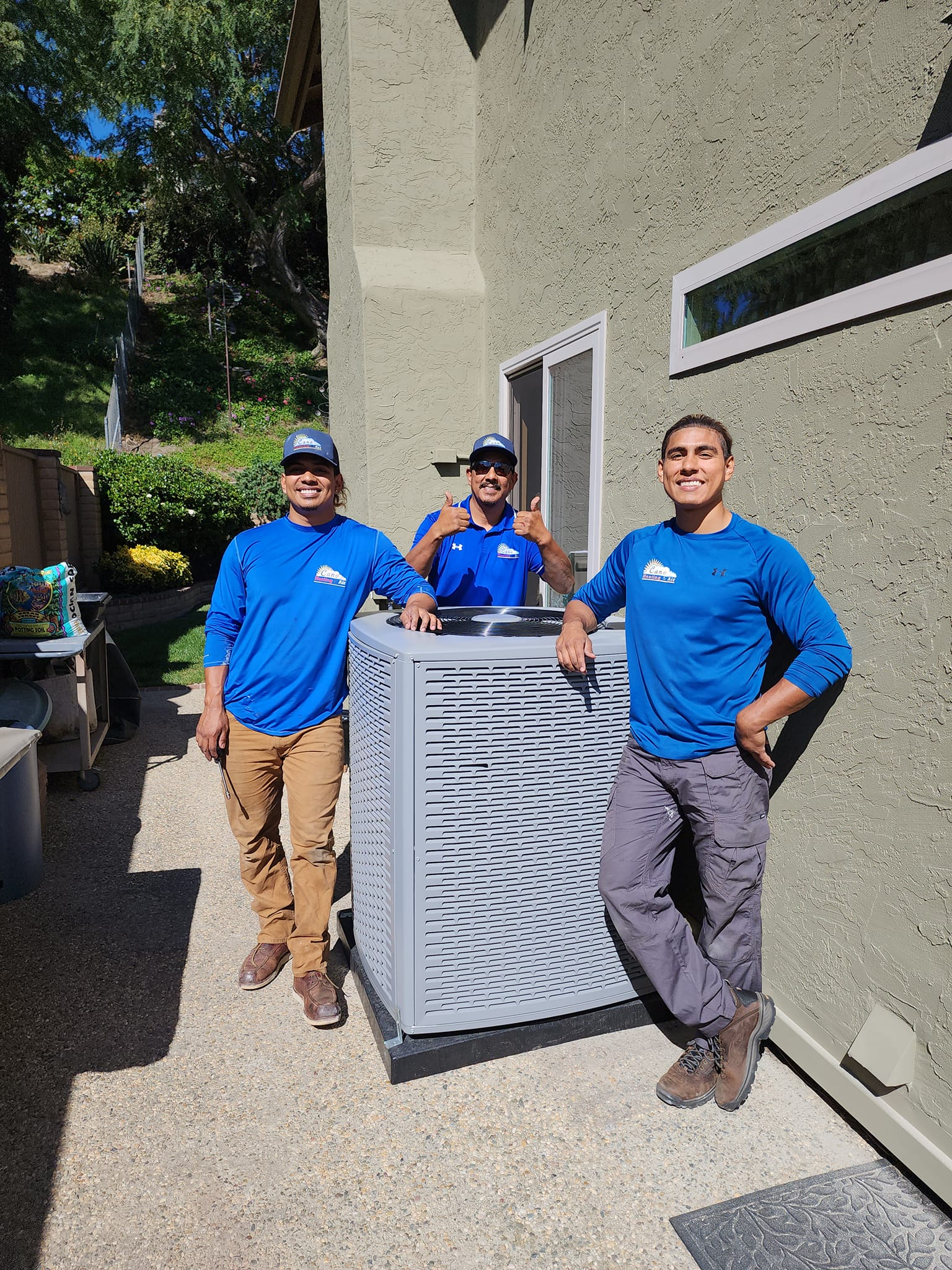 Three men in blue shirts stand with a new gray air conditioning unit outside a building.