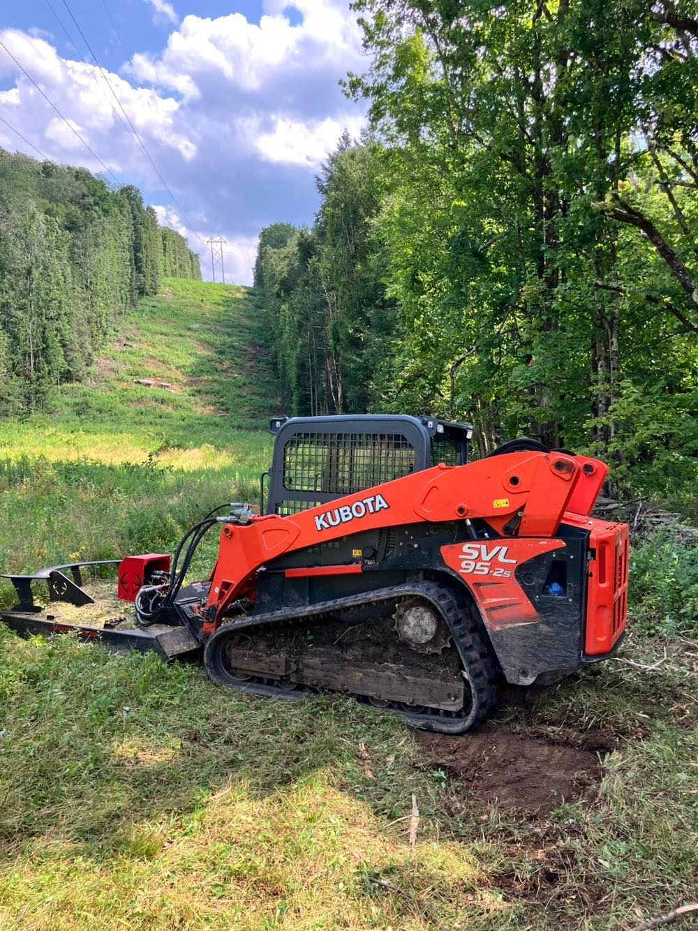 Orange Kubota skid steer with mower attachment on a grassy hillside beside a forested clearing