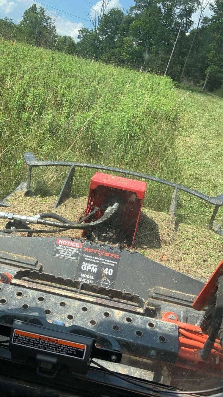 View from a red mower driving through tall green grass in a field, with trees in the background