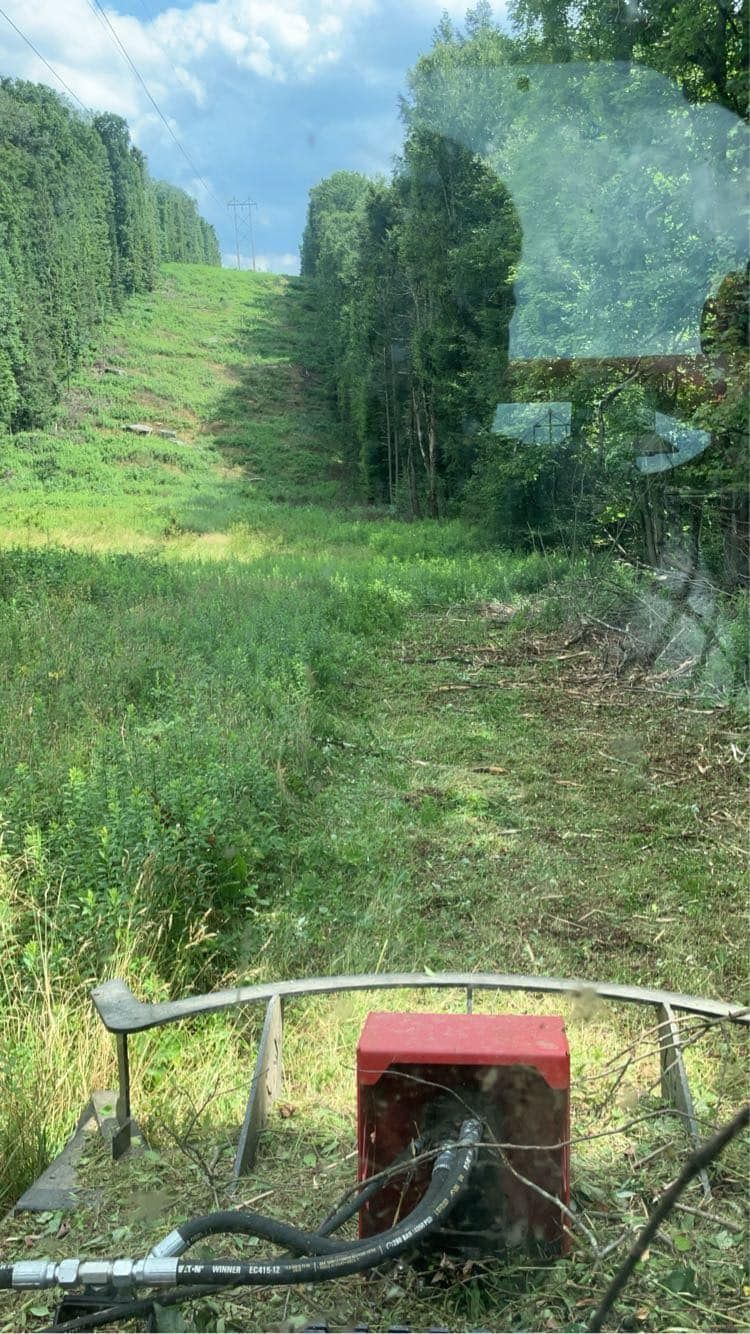 View from a lawn mower toward a grassy hillside path with trees and blue sky.