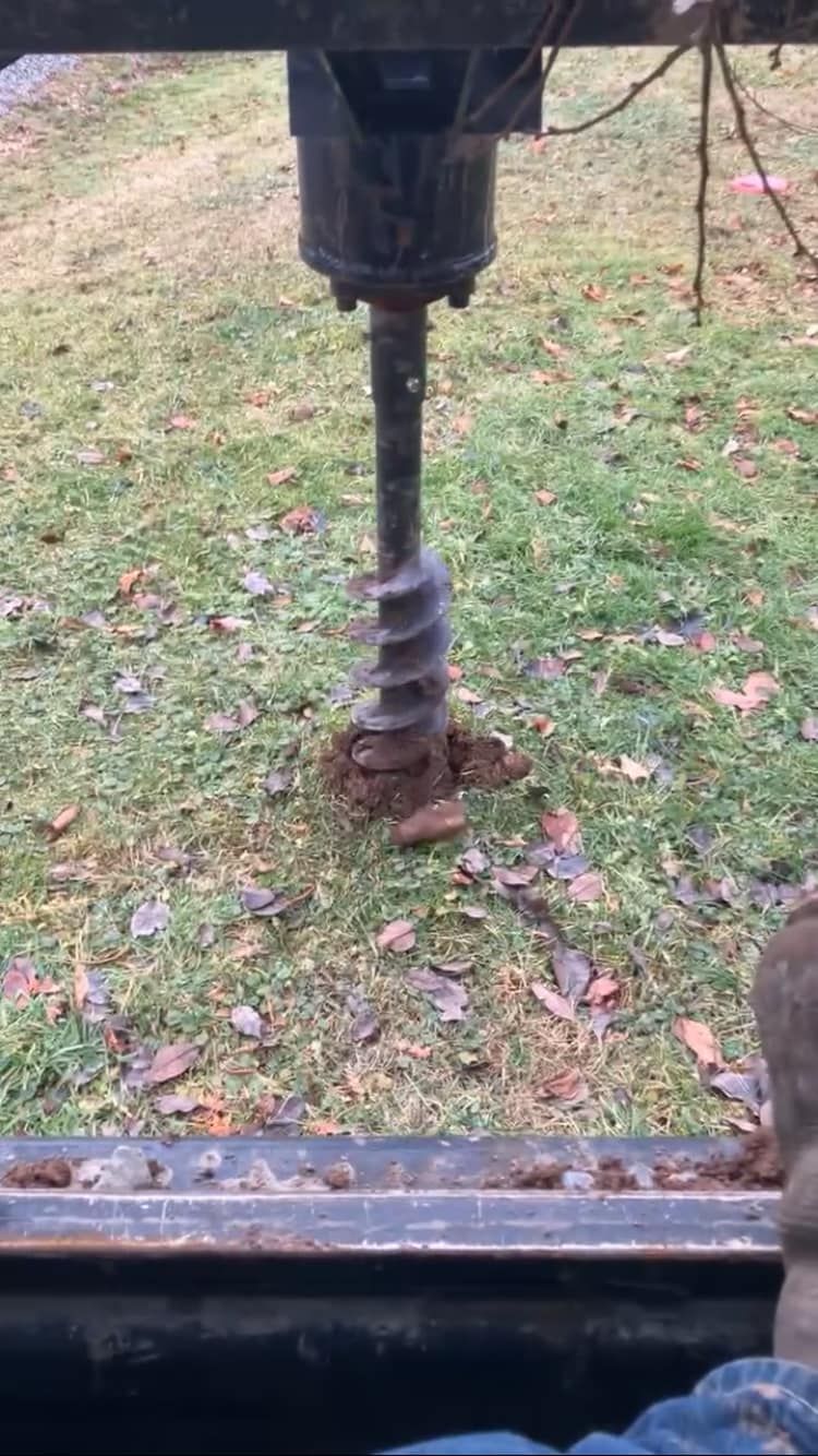 Rusty drill auger attached to machinery, boring into the ground on a grassy field.