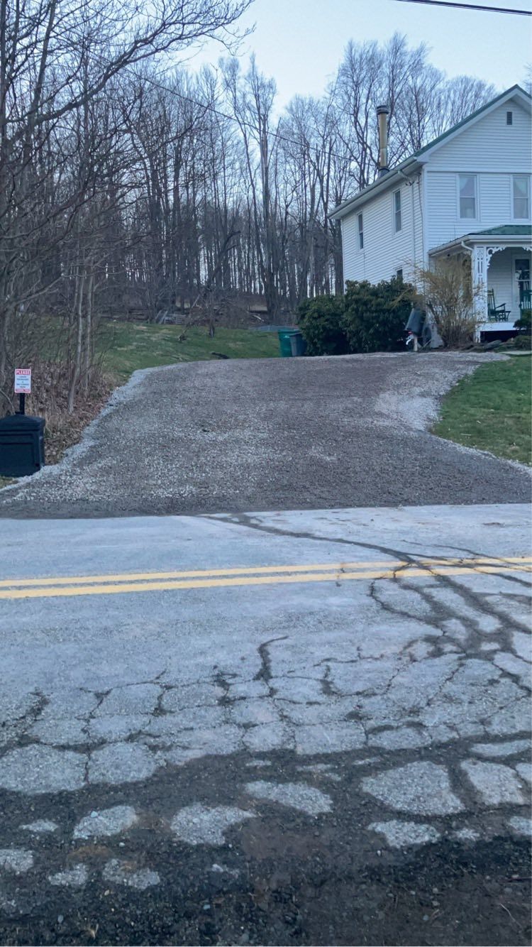 Driveway leading to a white house beside a wooded yard at dusk, with cracked pavement in the foreground