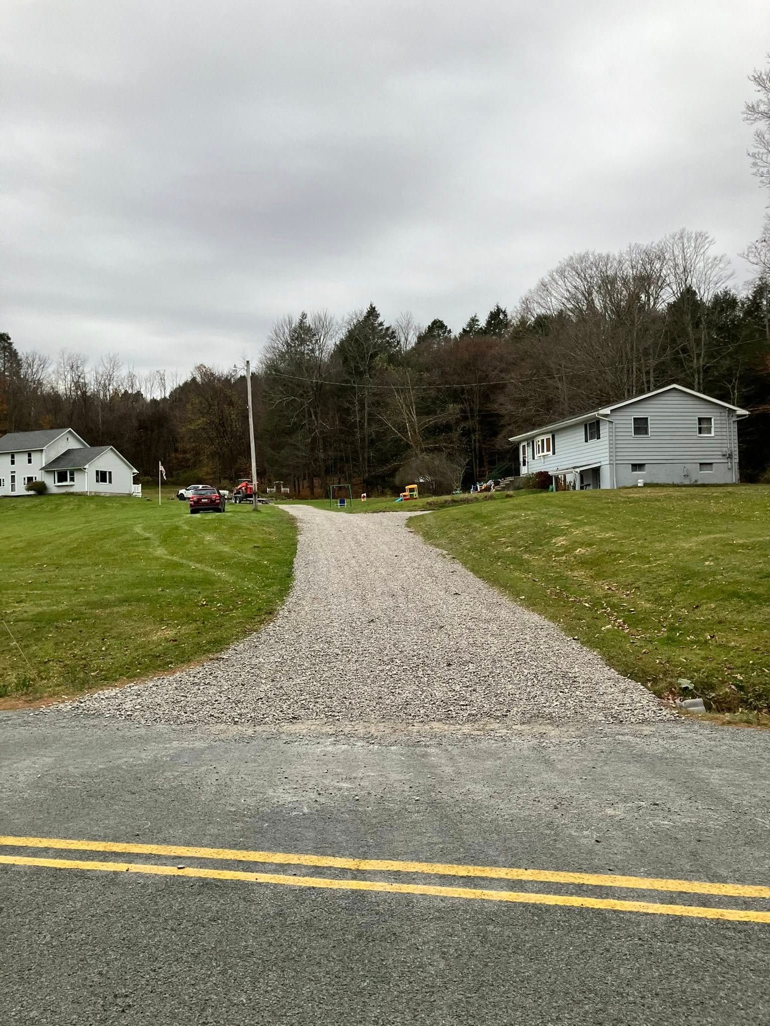 Gravel driveway leading to houses across a grassy field under a cloudy sky, with double yellow road lines in front.