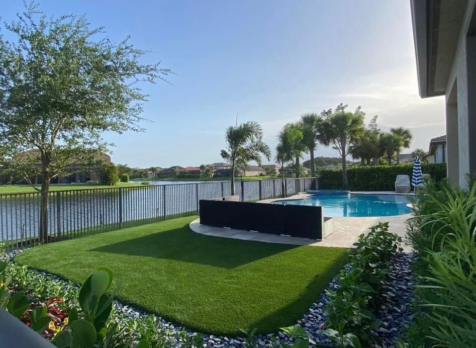 Lakeside backyard with a pool, green lawn, fence, and palm trees under a bright blue sky.