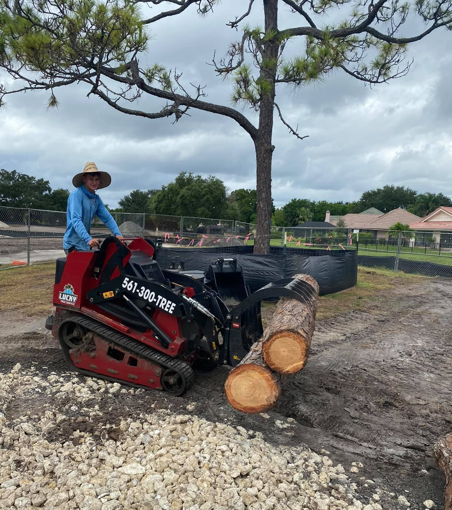 Worker on a red skid-steer loader moving cut tree logs in a muddy lot under cloudy skies