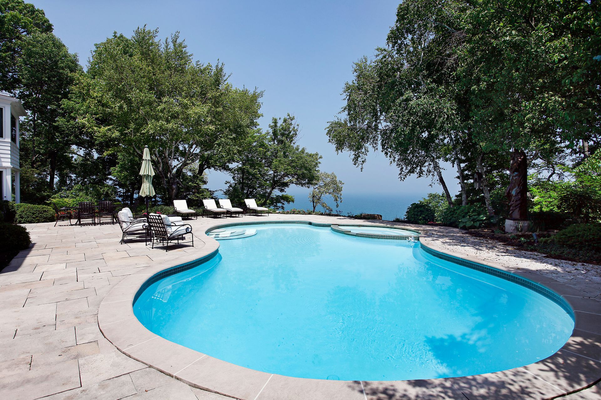 Curved turquoise pool surrounded by lounge chairs, trees, and a ocean view