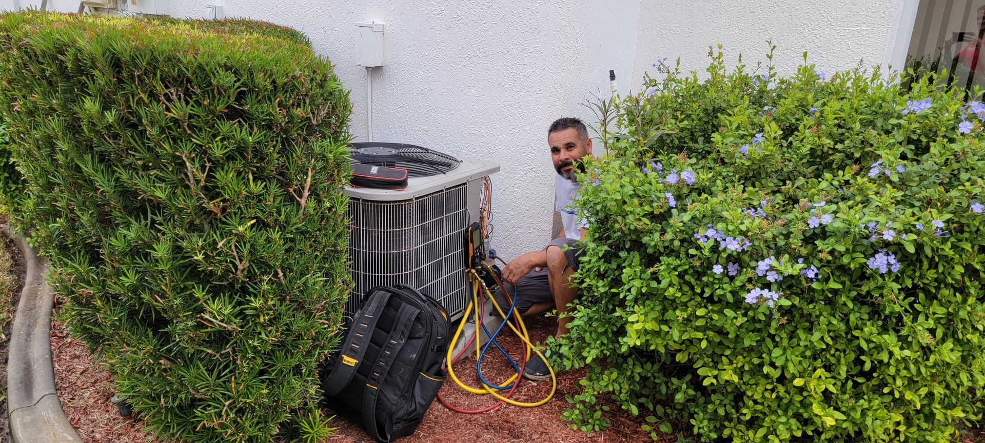 A person works on an air conditioning unit surrounded by bushes. There's a black toolbox on the ground.