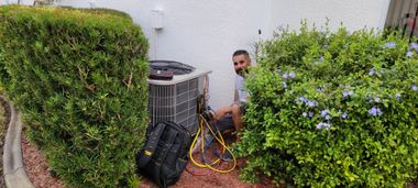A person works on an air conditioning unit surrounded by bushes. There's a black toolbox on the ground.