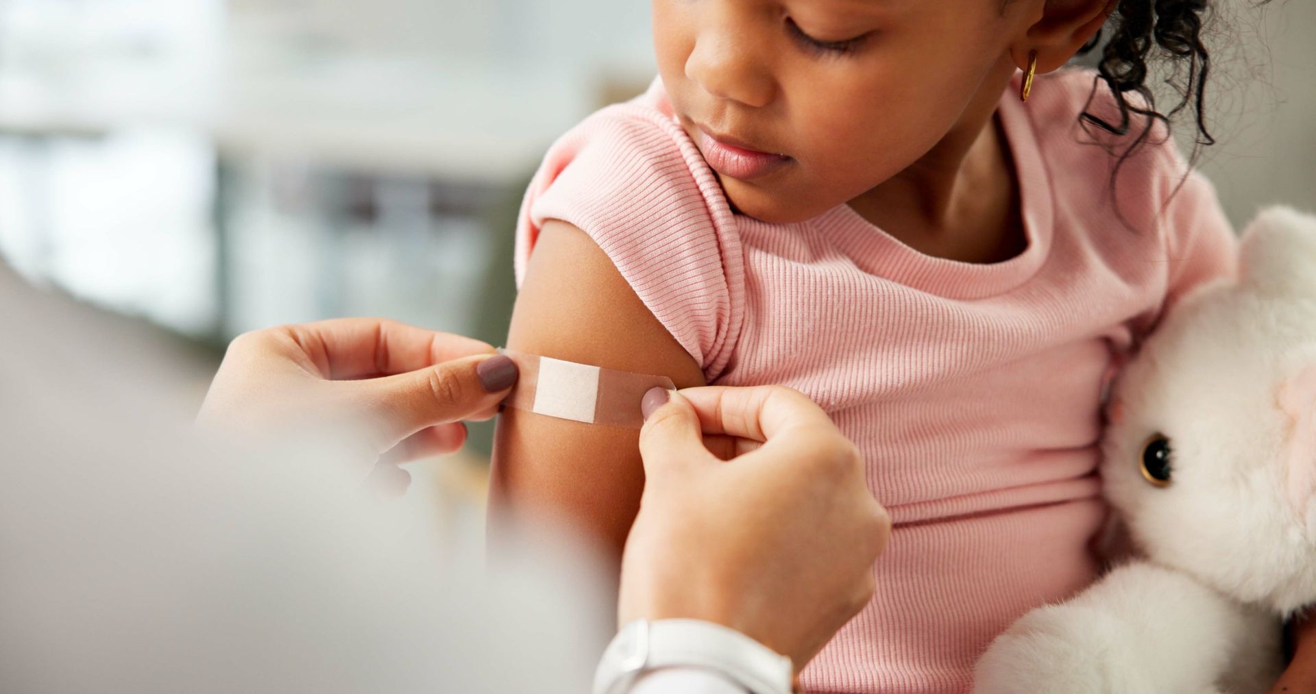 A caregiver applies a bandage to a child’s upper arm while the child holds a stuffed rabbit.