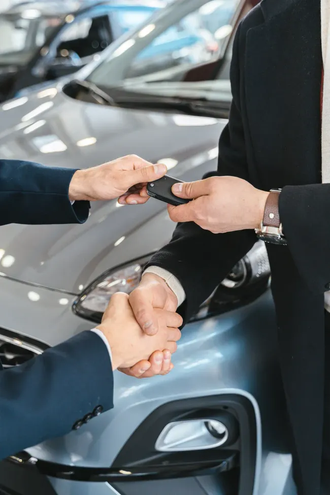 Hands exchanging car keys in a dealership showroom beside a gray car.