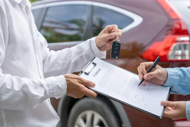 Hands exchanging a car key and signing paperwork beside a red vehicle