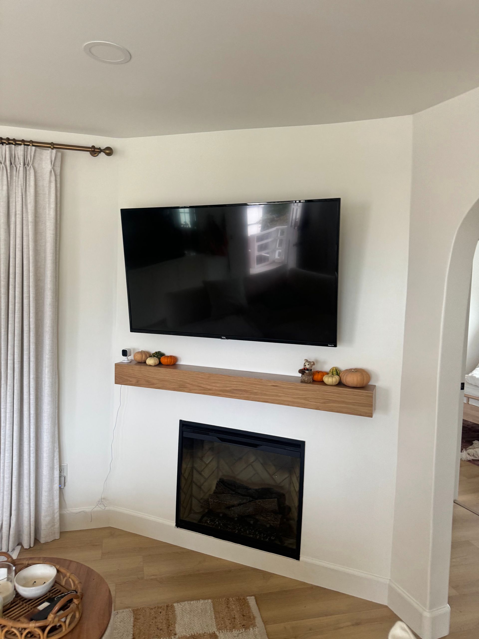Living room corner with mounted TV above a white fireplace and wooden mantel, beside sheer curtains.
