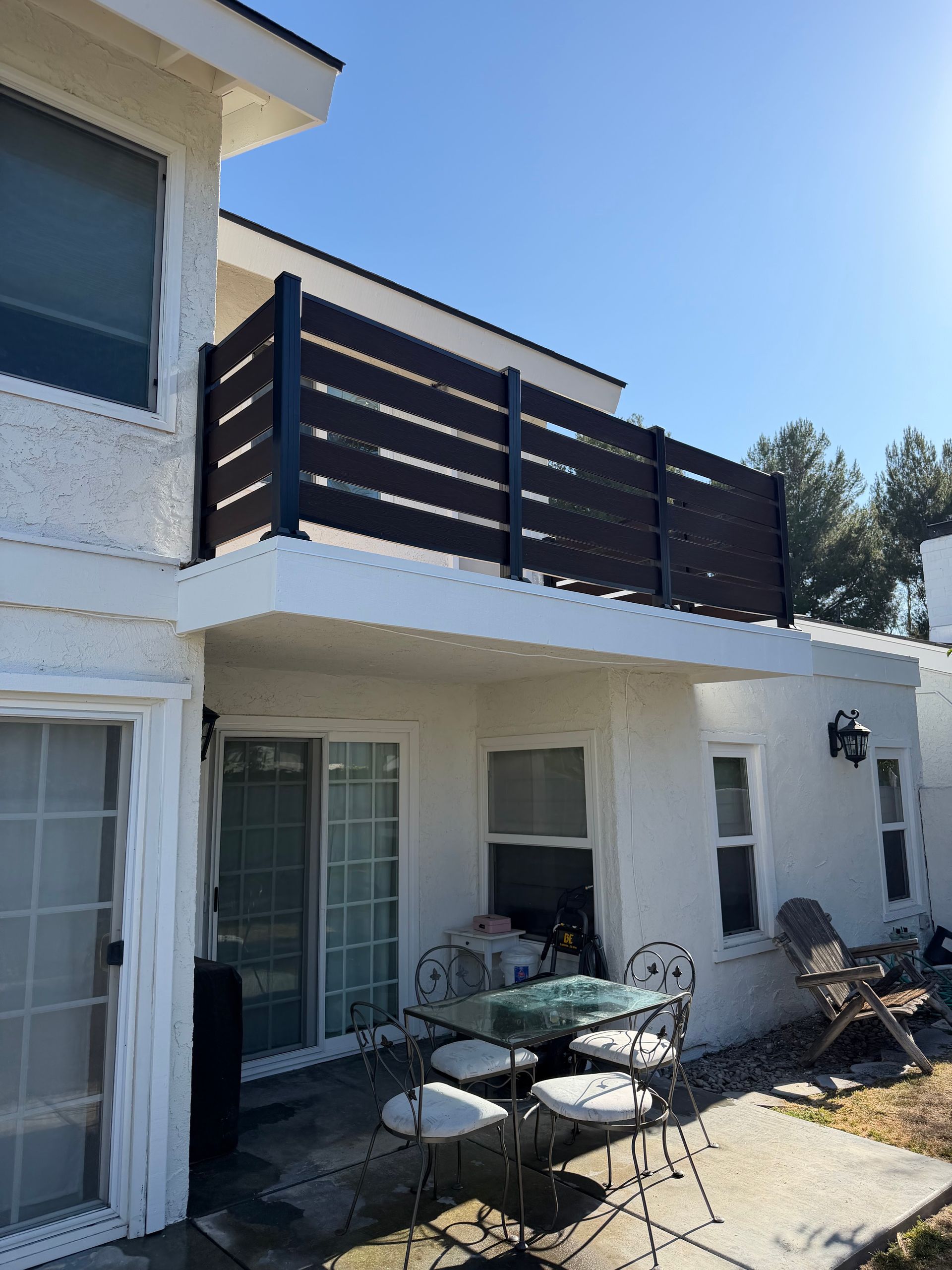 Patio with glass table and chairs beside a two-story house with a balcony and dark privacy slats