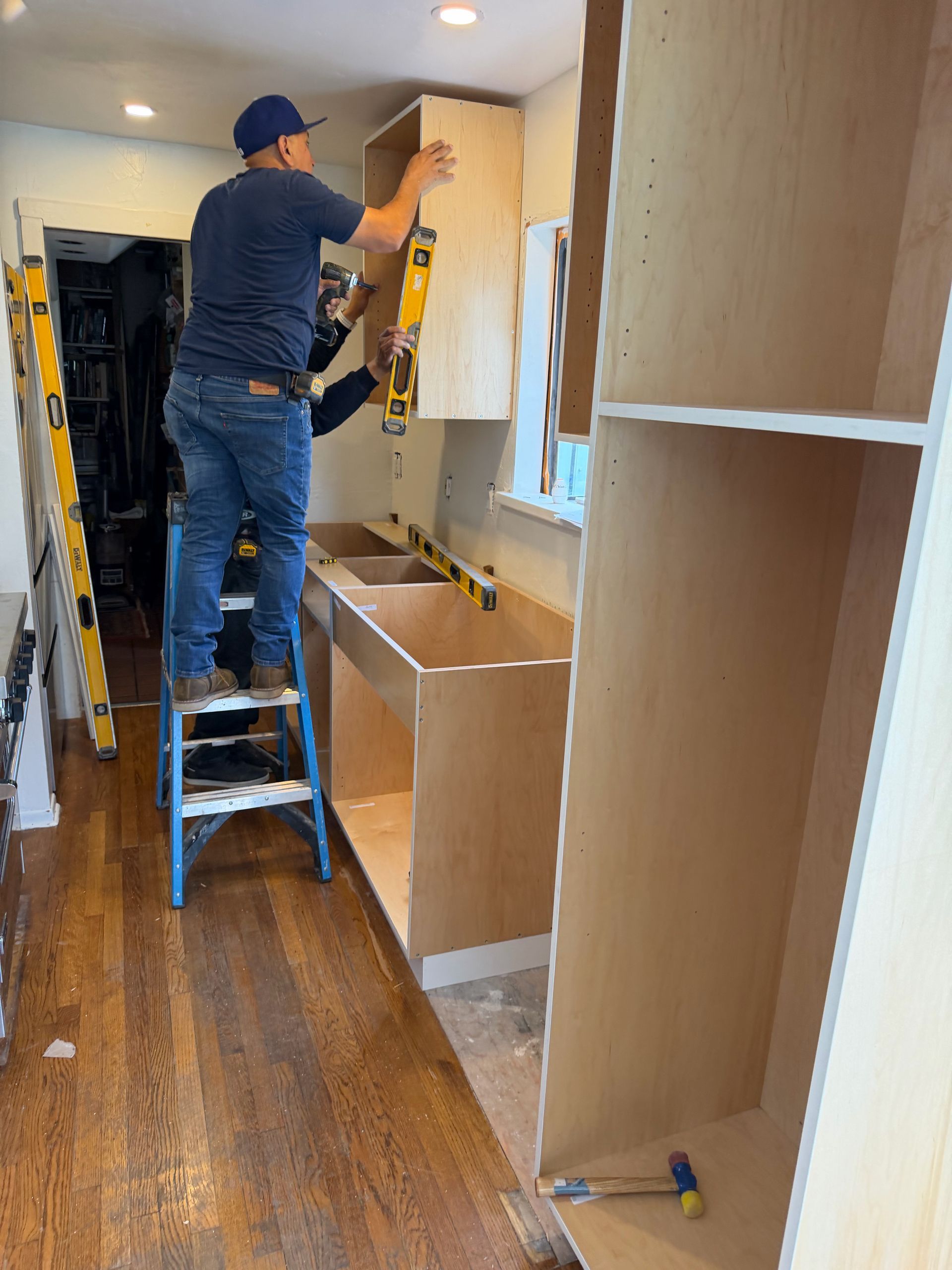 Worker installing cabinets in a narrow hallway, standing on a step stool with tools and unfinished cabinetry.