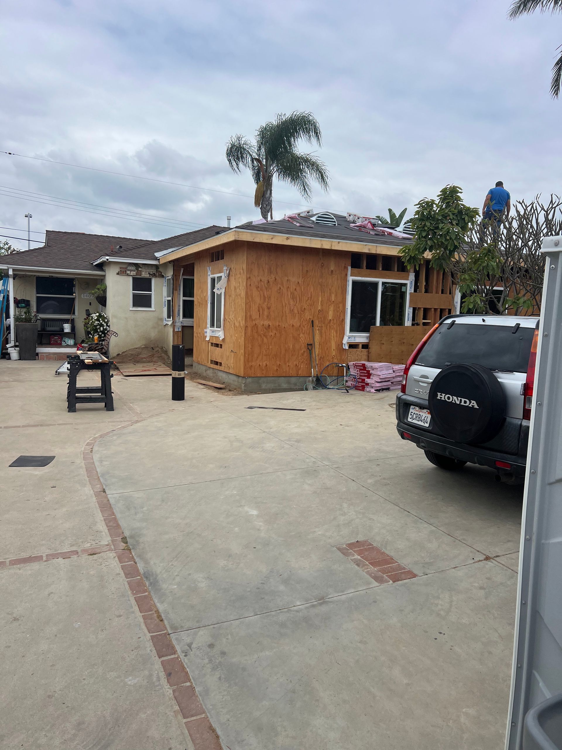 Courtyard with a small wooden house, parked SUV, and palm trees under a cloudy sky.