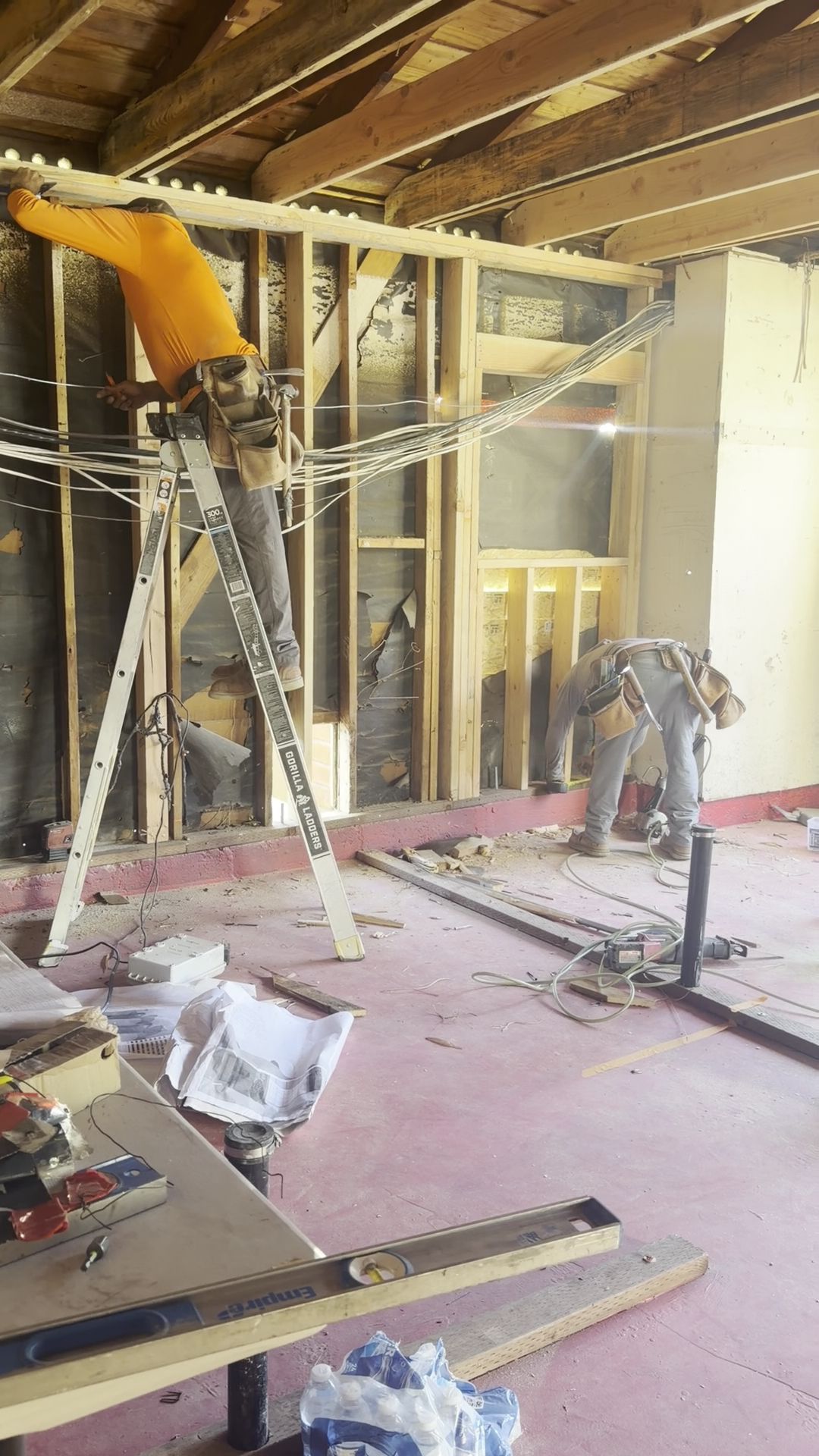 Worker on a ladder in a room under renovation, with exposed walls, beams, and construction materials.