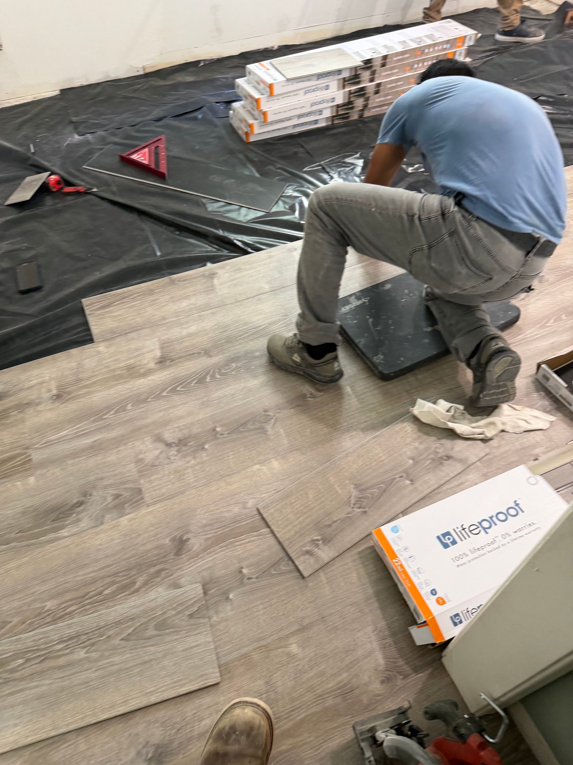 Worker kneeling on a partially installed wood floor beside black underlayment and boxes of materials