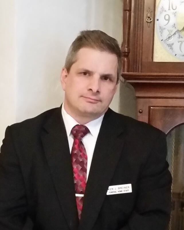 Man in a black suit and red tie standing indoors beside a wooden cabinet and wall clock.