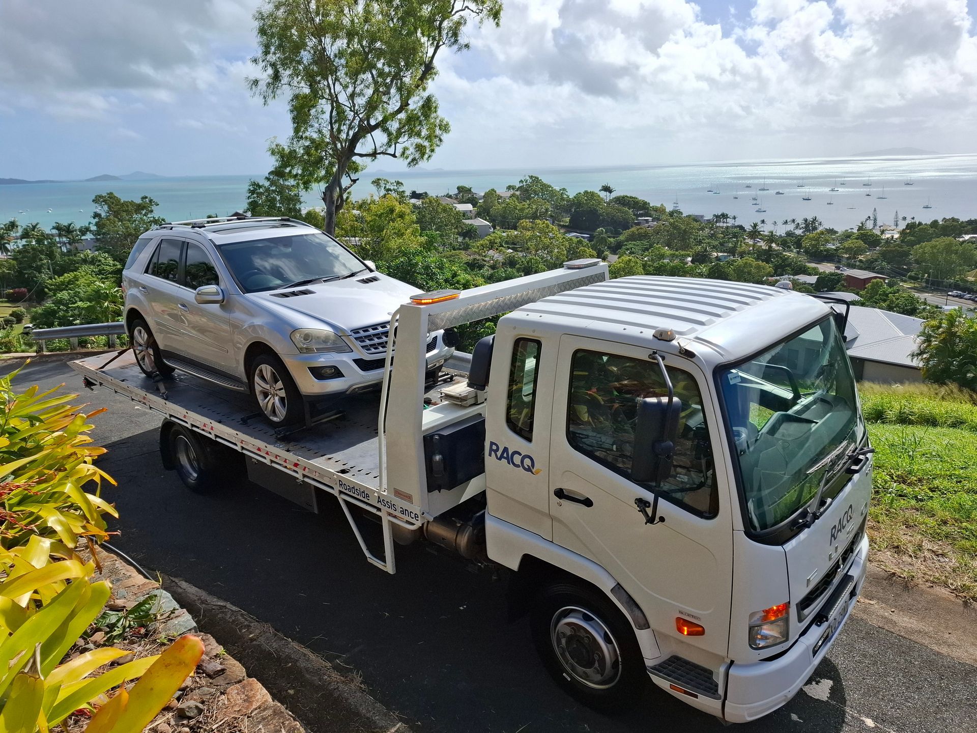 Tow truck carrying a silver SUV on a hillside road with ocean view in the background — Airlie Towing & Tilt Tray Hire In Cannonvale, QLD