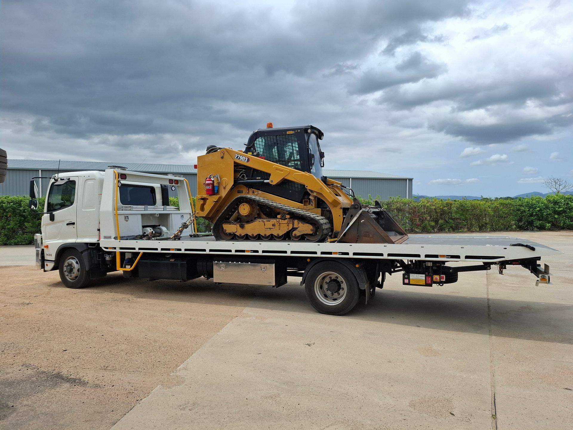 White Flatbed Truck Carrying a Yellow Tracked Loader on a Roadside Under Cloudy Skies — Airlie Towing & Tilt Tray Hire In Cannonvale, QLD