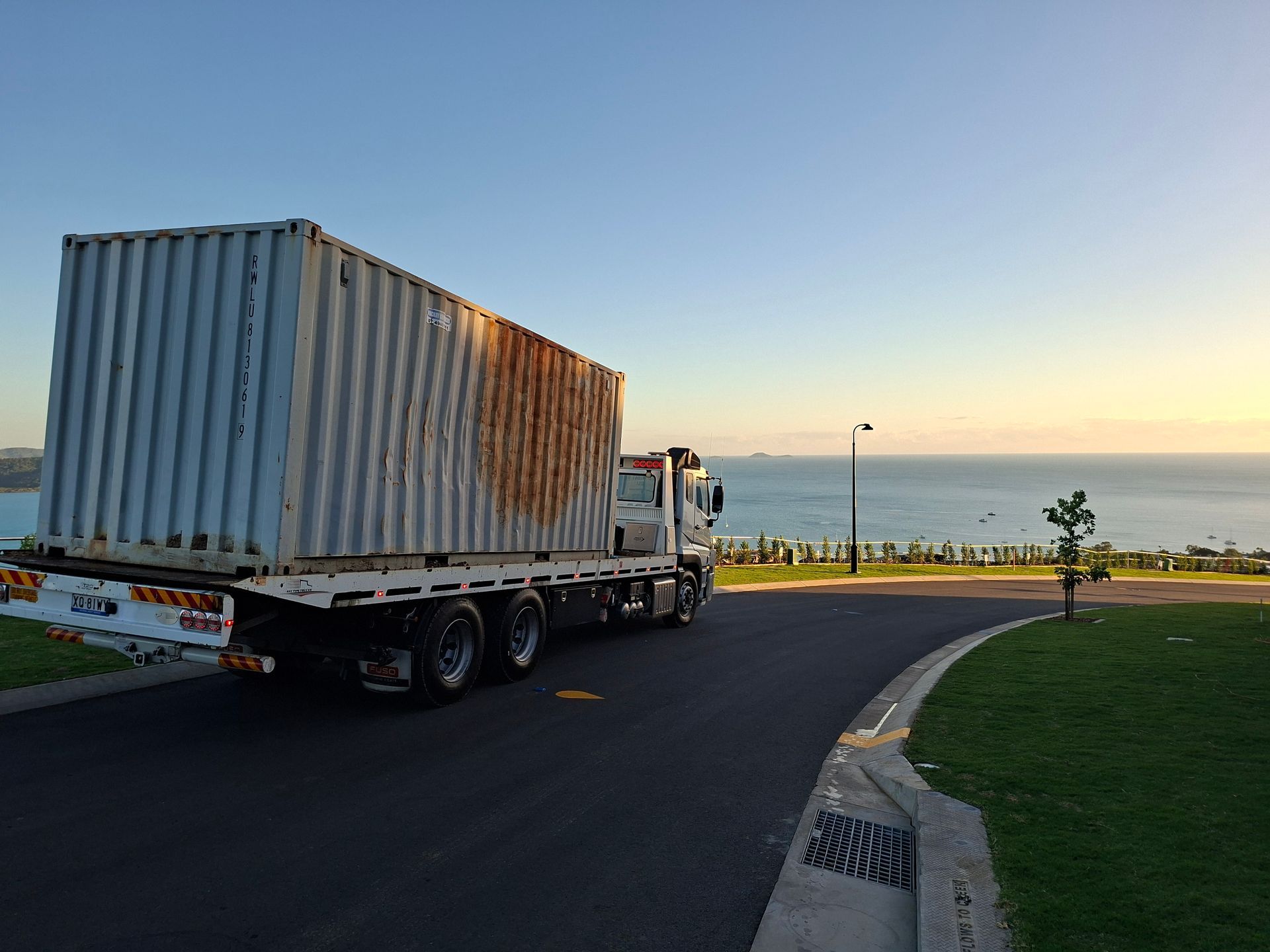 White shipping container on a flatbed truck driving by the ocean at sunset— Airlie Towing & Tilt Tray Hire In Bowen, QLD