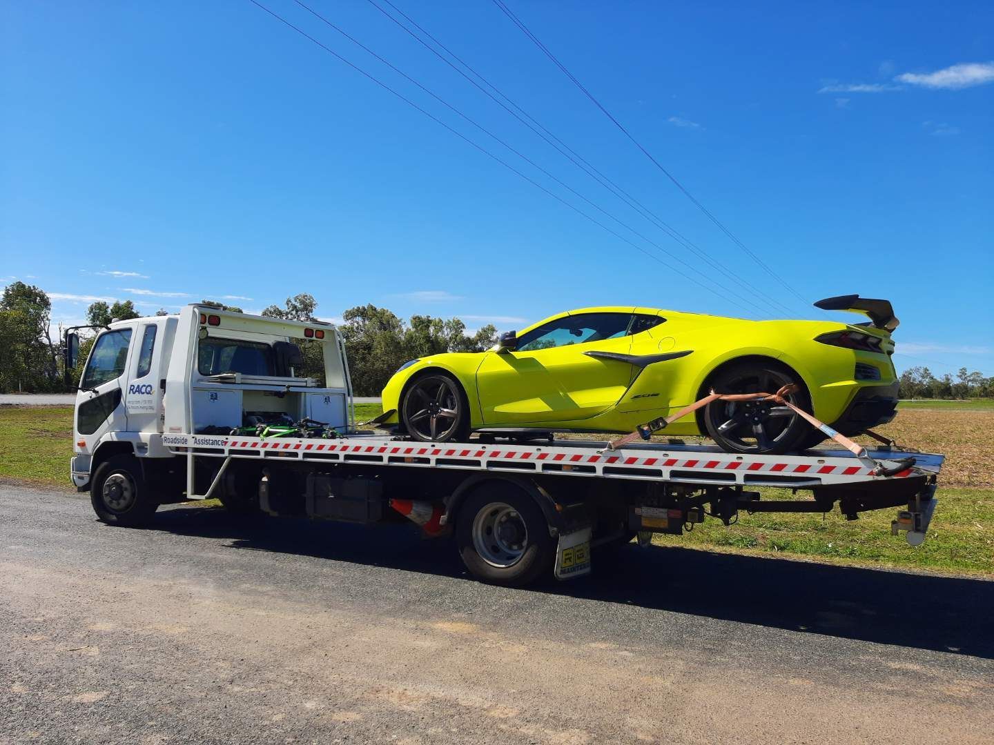 Tow Truck Carrying a Bright Yellow Sports Car on a Flatbed Along a Roadside Under a Clear Blue Sky — Airlie Towing & Tilt Tray Hire In Cannonvale, QLD