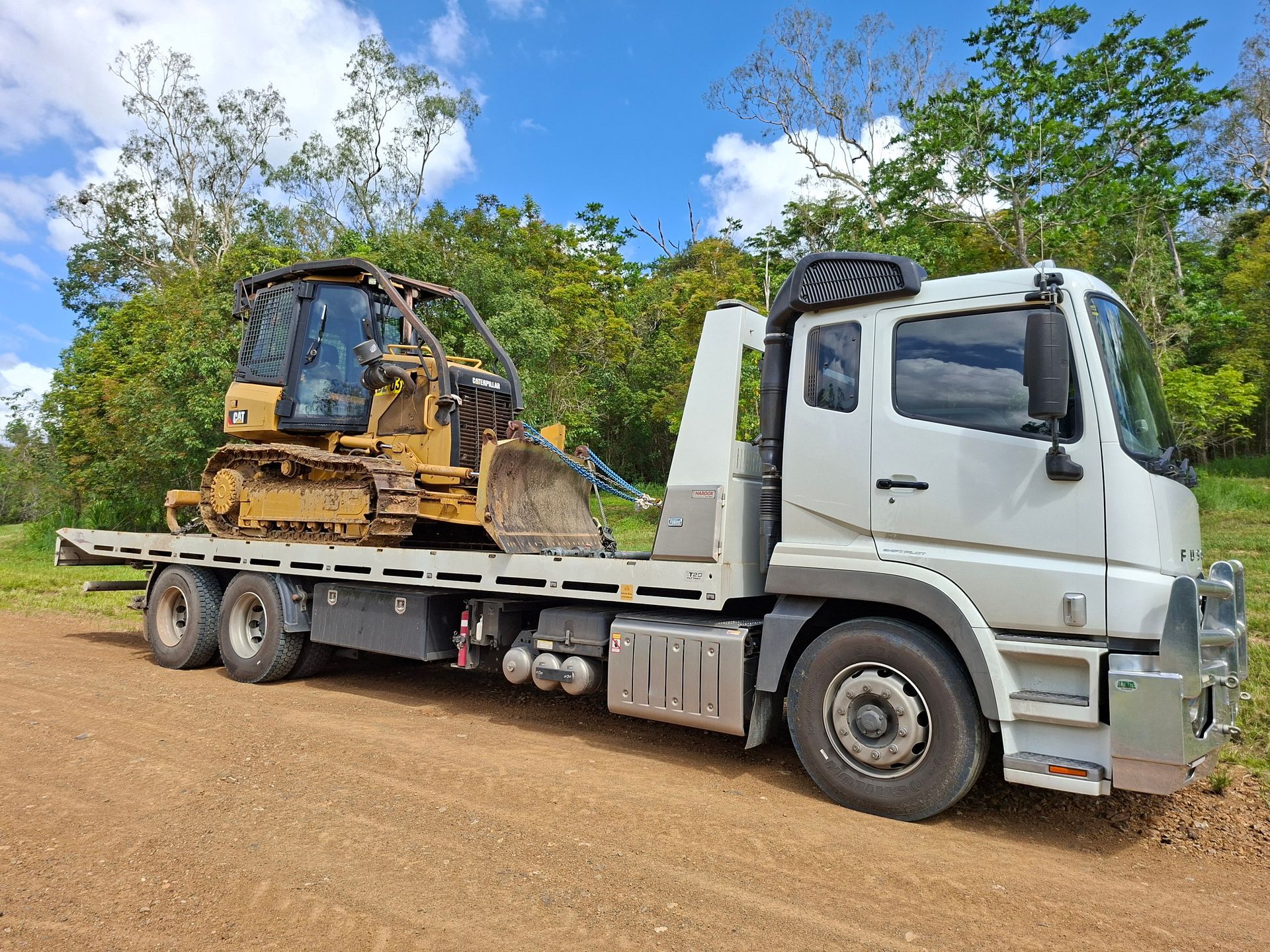 White Flatbed Truck Carrying a Yellow Bulldozer on a Dirt Road With Trees and Blue Sky — Airlie Towing & Tilt Tray Hire In Cannonvale, QLD