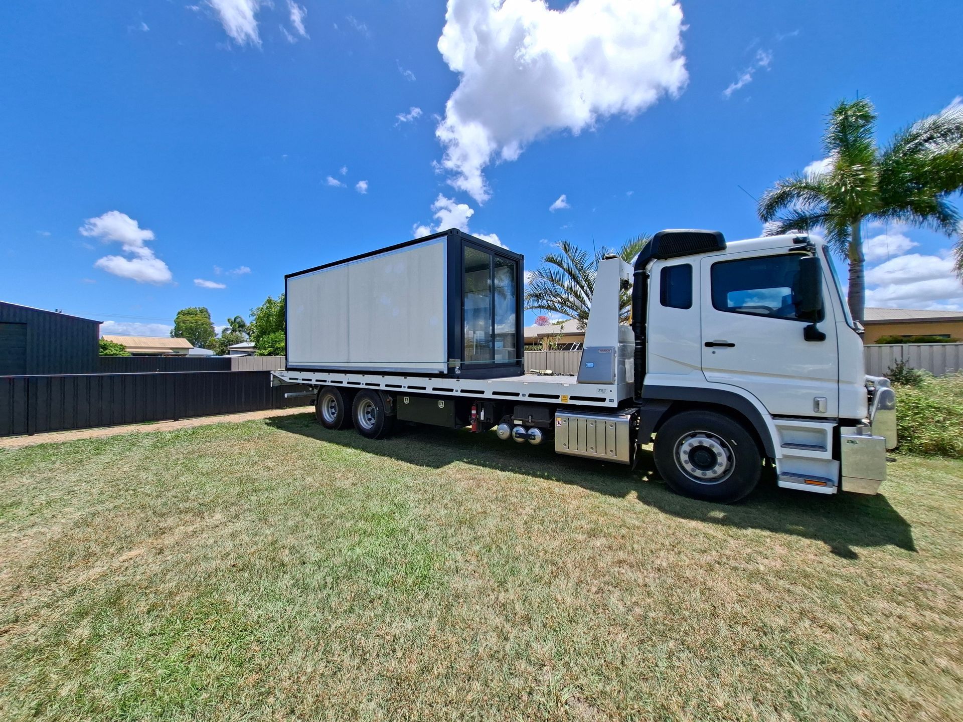 White Flatbed Truck Hauling a Gray Container on a Grassy Roadside Under a Blue Sky — Airlie Towing & Tilt Tray Hire In Cannonvale, QLD