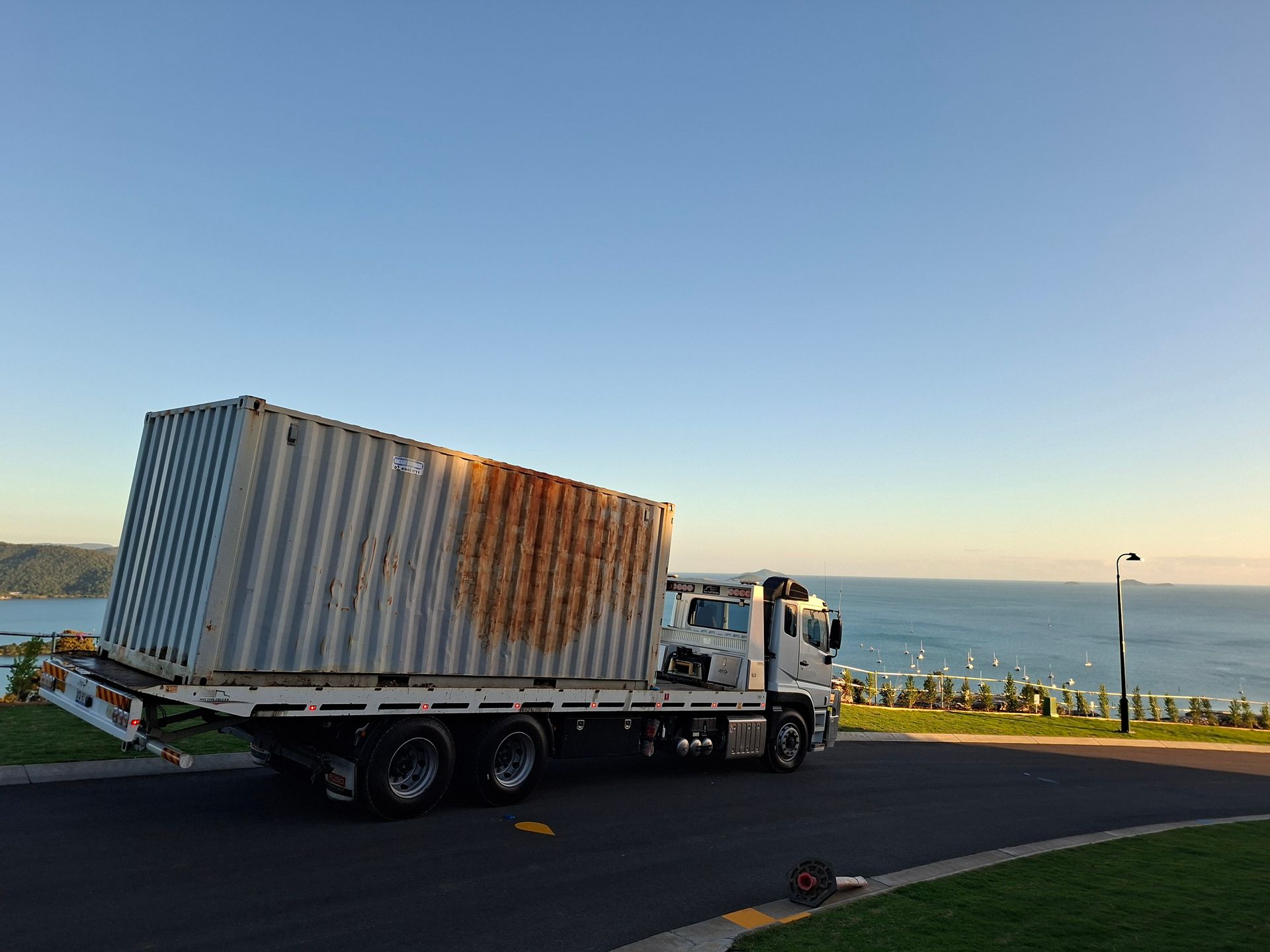 White Flatbed Truck With a Rusty Shipping Container Parked by the Waterfront at Sunset — Airlie Towing & Tilt Tray Hire In Airlie Beach, QLD