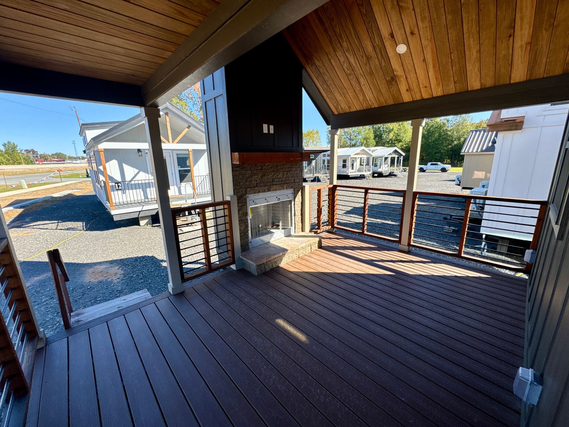 Covered wooden porch with a stone fireplace and railing overlooking a gravel lot with several manufactured homes.