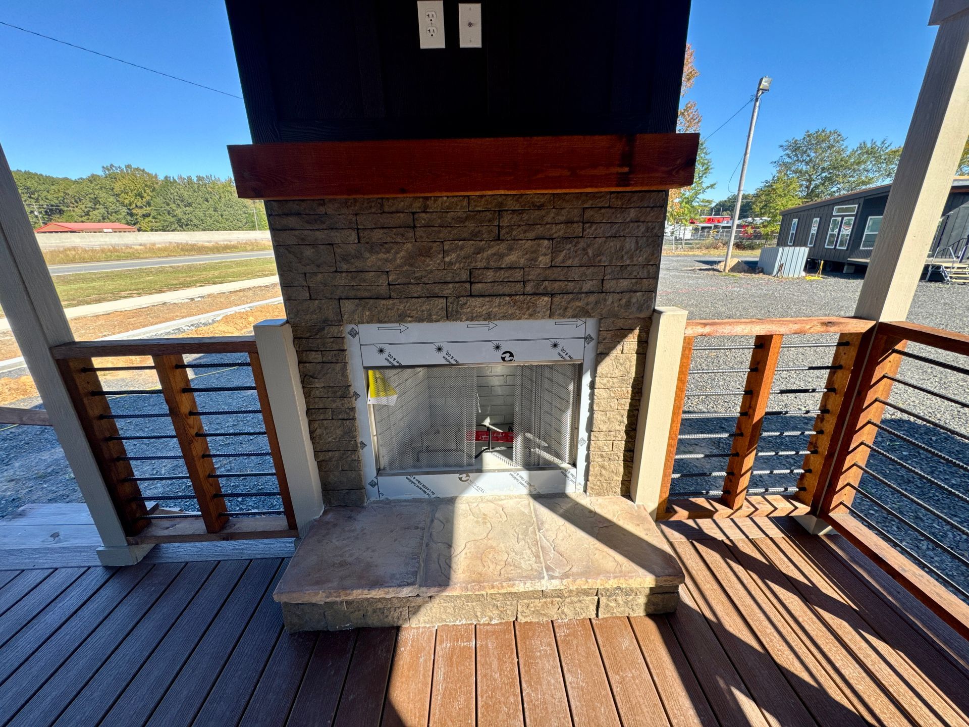 Outdoor stone fireplace on a wooden deck with railing, featuring a wooden mantel and a protective plastic window cover.