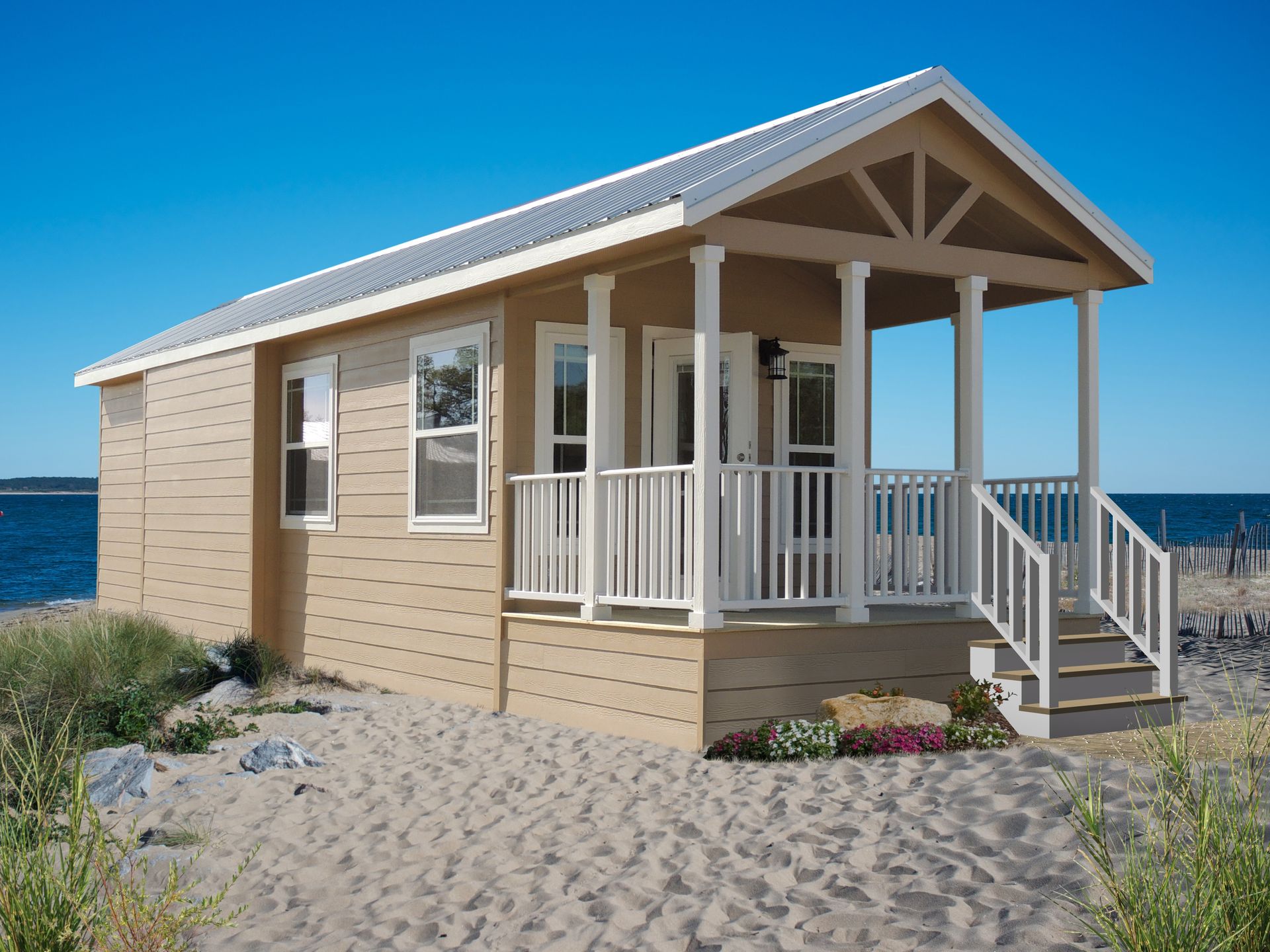 Tan beach cabin with porch and steps, white railings, metal roof, on sand. Blue sky and water in the background.