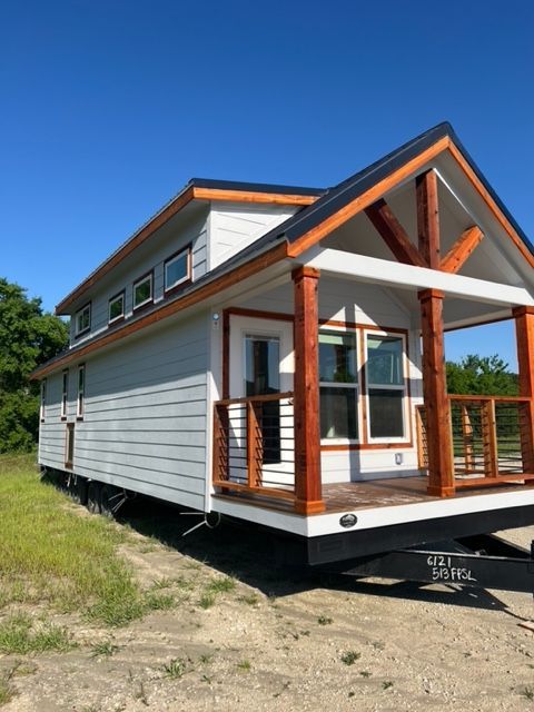 Tiny house on a trailer with white siding, wood accents, and porch. Bright sunny day.