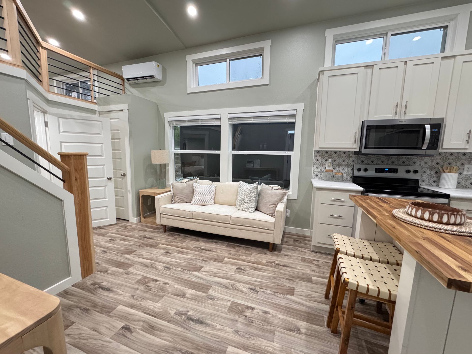 A light-colored living room with a sofa, a wooden bar with two stools, a kitchen area, and a loft area with a railing.