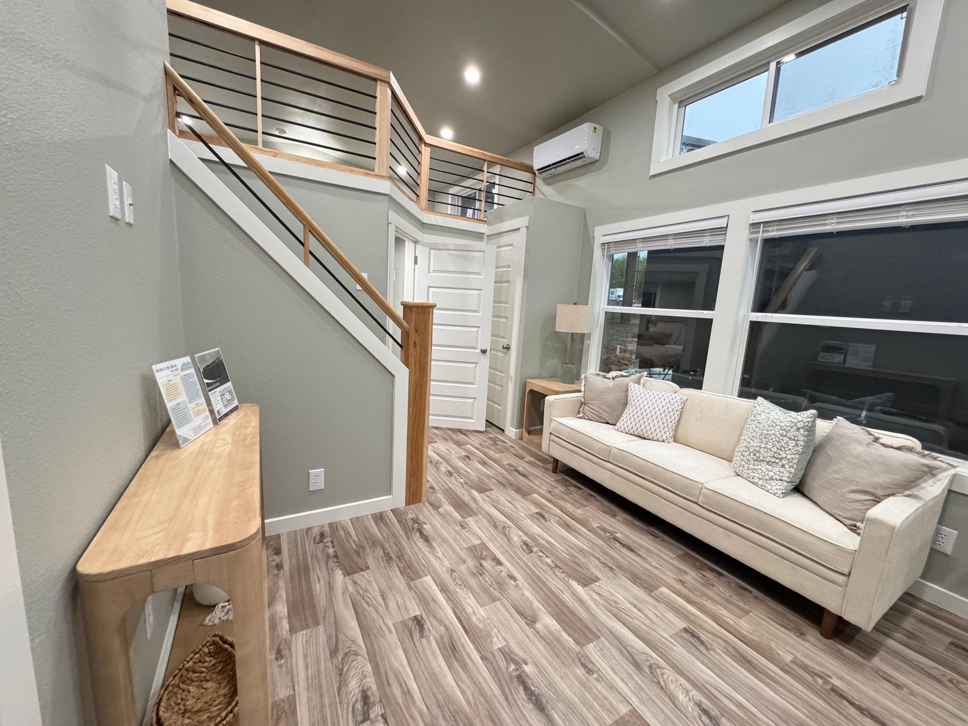 A modern tiny home living room with a beige sofa, wood-look flooring, a loft area with railings, and a console table.
