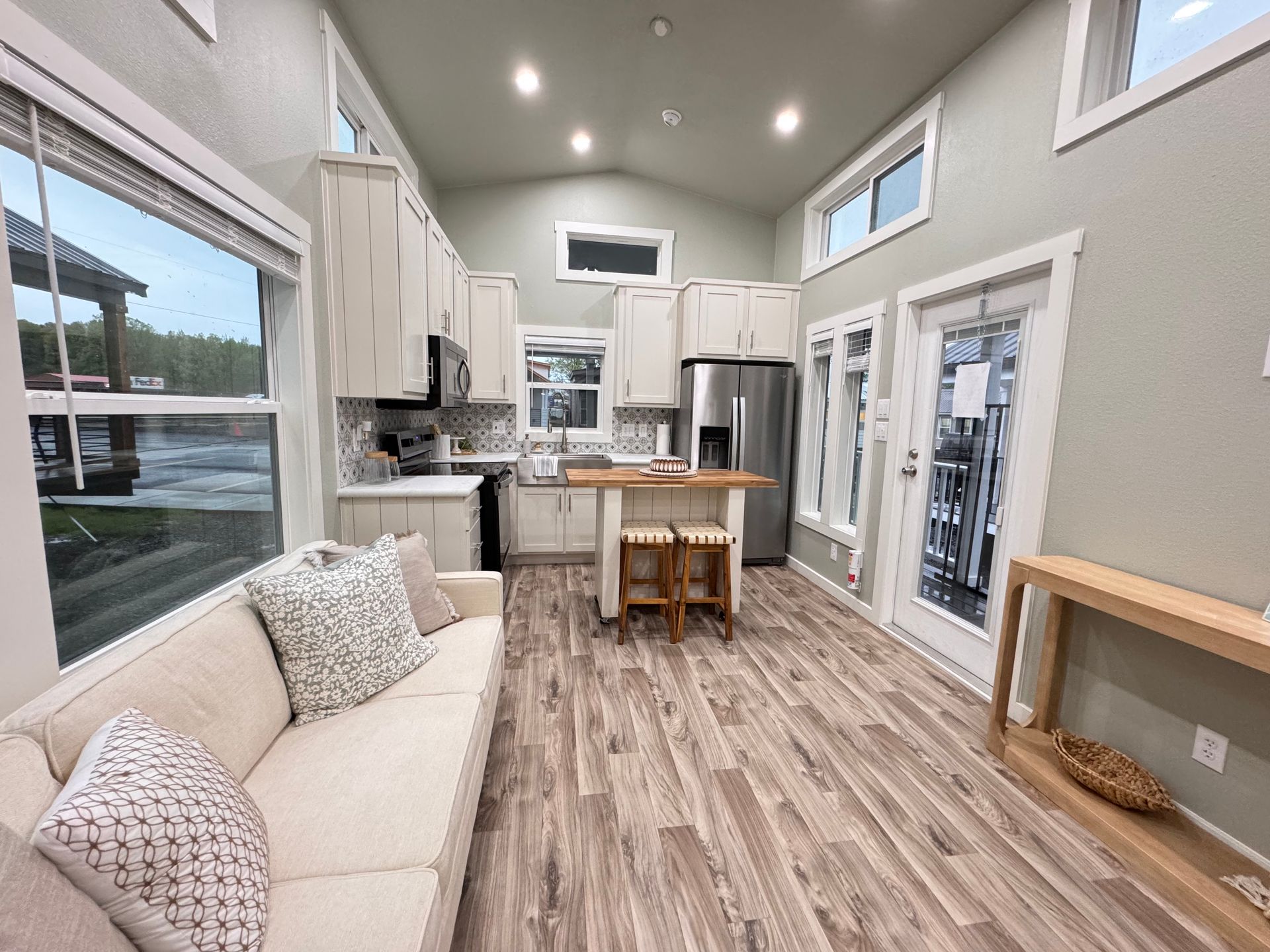 A bright tiny home interior features a light-colored sofa, wood-look floors, a small kitchen island, and stainless fridge.