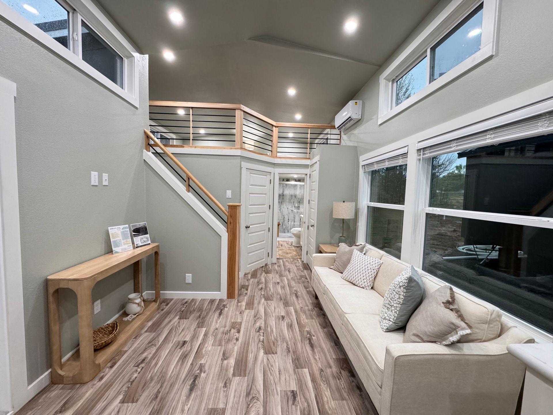 Modern tiny home interior featuring a white sofa, wood-look floors, a wooden console table, and a loft with railing.